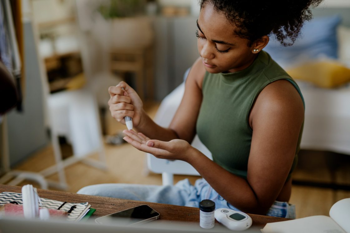 Teenage girl collecting fingerstick blood sample from finger for blood sugar testing at home, during day. Student girl testing blood sugar levels with a glucometer. Paediatric diabetes in teenage girl and life with chronic illness.