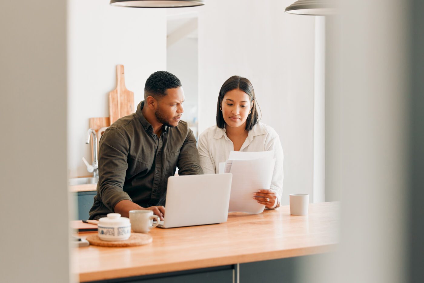 A man and woman standing in a kitchen looking over papers and a laptop.