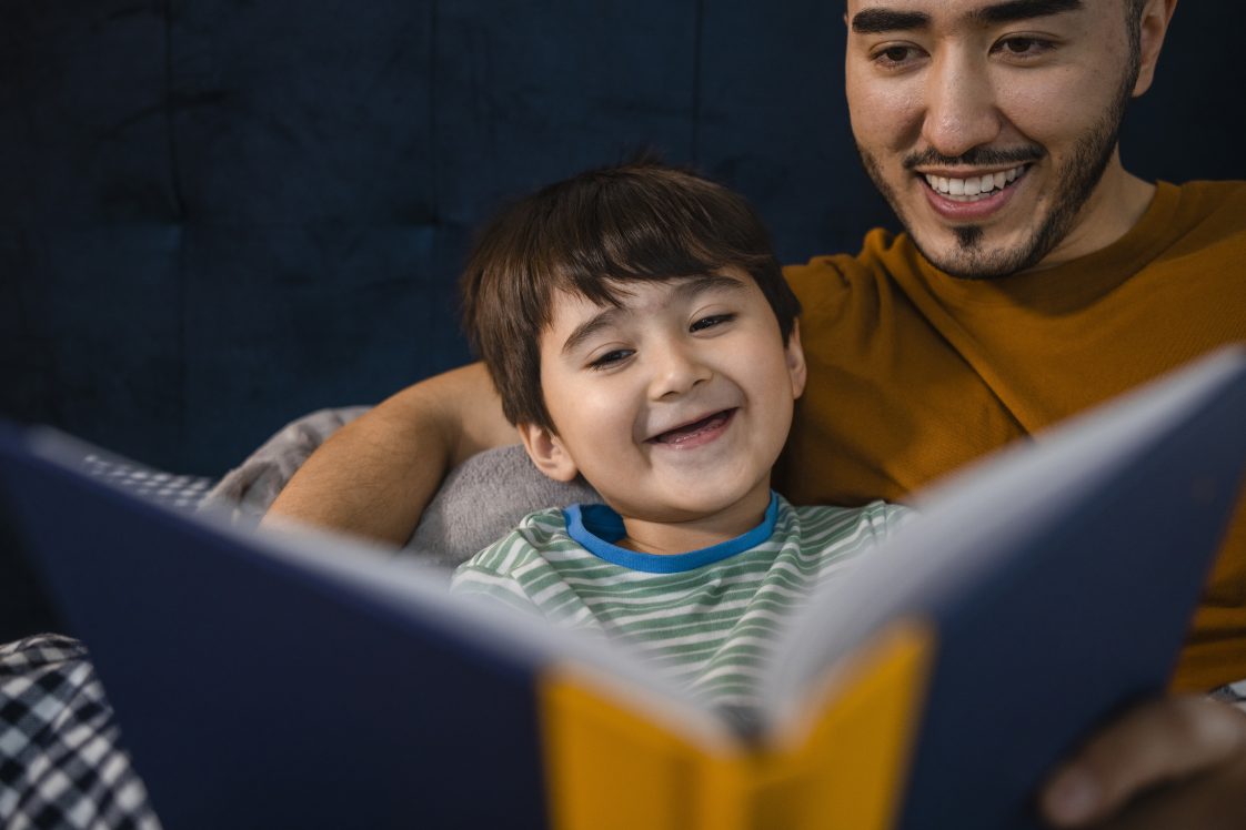An Asian man and his young son wearing pajamas in his bedroom on a winter's night. They are lying in bed while the father reads his son a bedtime story.