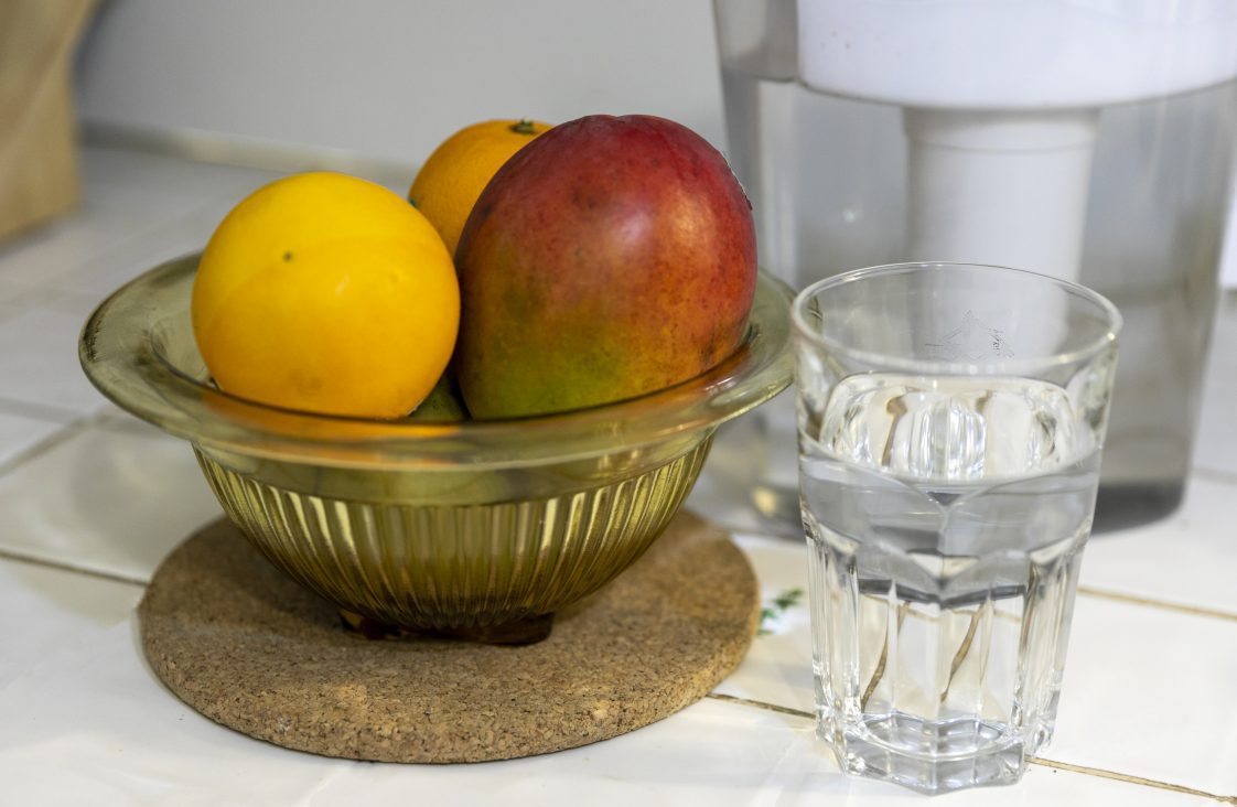 A fruit bowl with two oranges and a mango and a glass of water sitting on a kitchen counter.