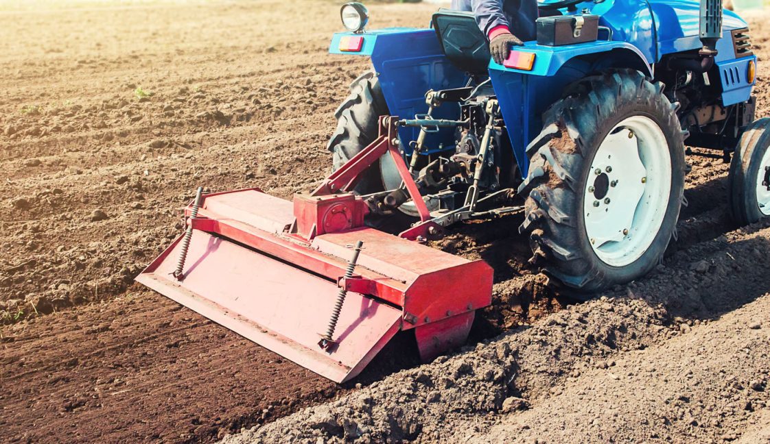 A blue tractor pulling a red tiller in a garden.