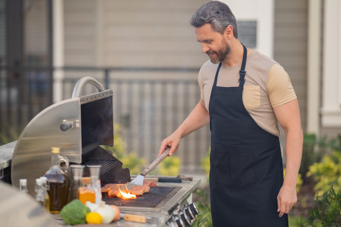 Man standing at grill using a metal spatula to flip hamburgers.