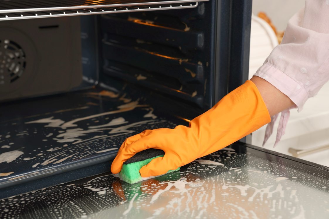 A woman's hand wearing an orange rubber glove and using a sponge to clean the oven.