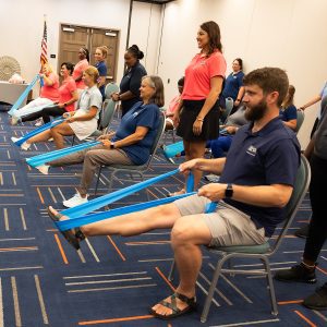 A room of adults doing seated resistance-band exercises.
