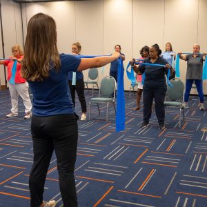 A room of adults being instructed doing resistance-band exercises.