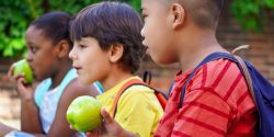 Multiethnic children with backpacks sitting on the street at school entrance eating apples and talking.