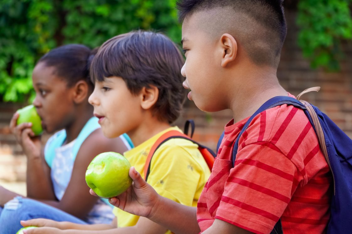 Multiethnic children with backpacks sitting on the street at school entrance eating apples and talking.