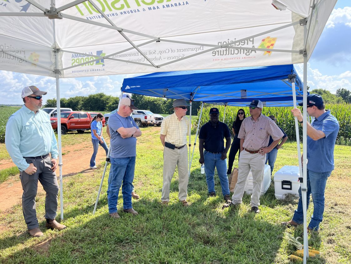 A group of people standing underneath a tent outside at a crop field day.