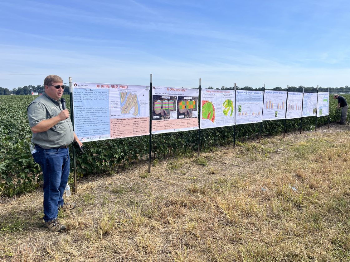 A speaker standing near the edge of a cotton field talking to people at a crops field day.