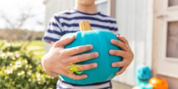 A young boy holding a teal pumpkin standing on a porch with other teal pumpkins in the background.