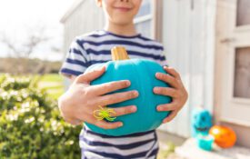 A young boy holding a teal pumpkin standing on a porch with other teal pumpkins in the background.