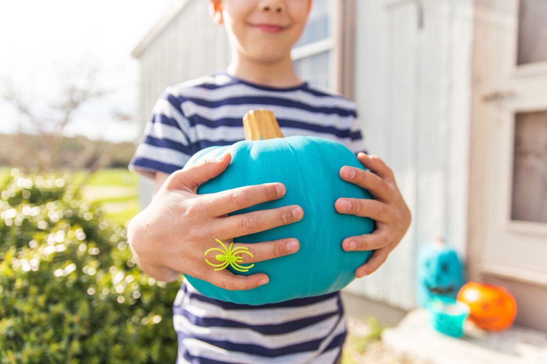 A young boy holding a teal pumpkin standing on a porch with other teal pumpkins in the background.