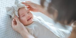 A closeup of a happy, smiling baby wearing a bow while lying on a bed.