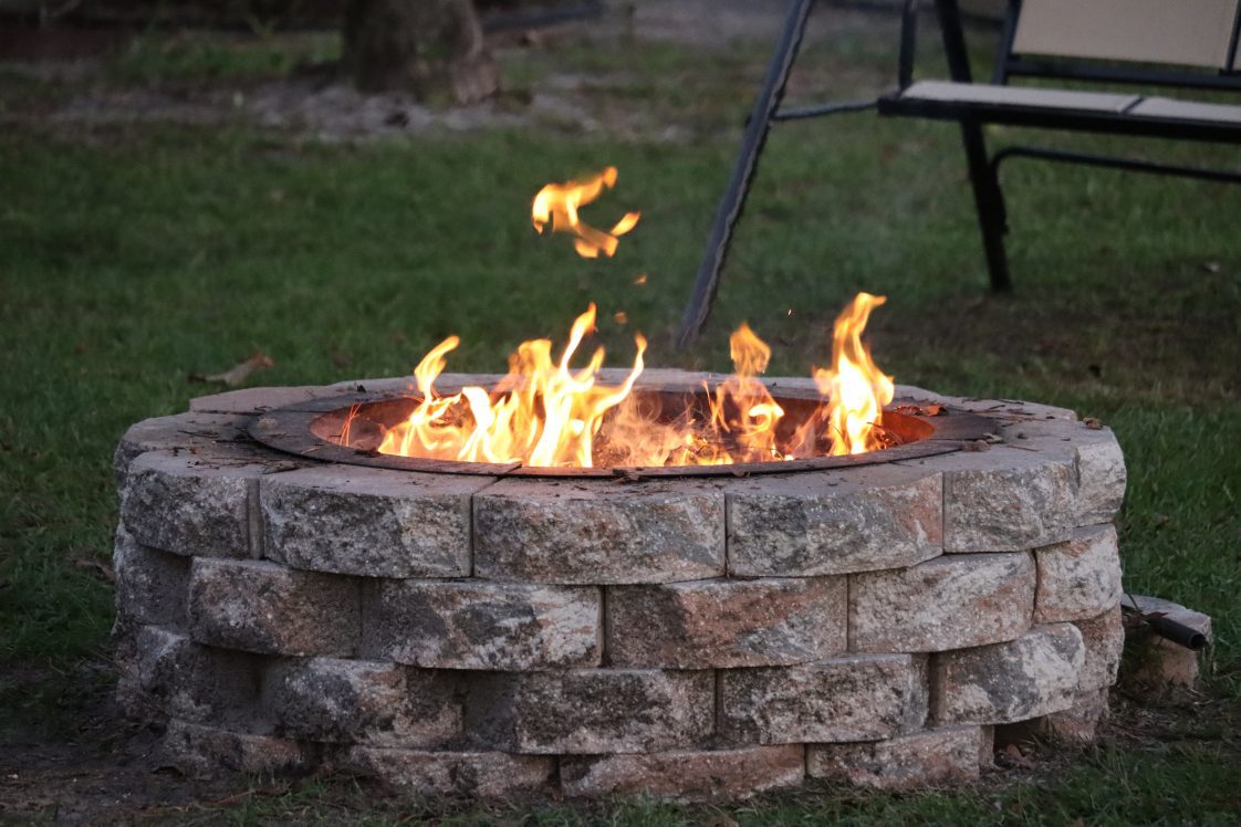 A stone fire pit with a light fire in a backyard.