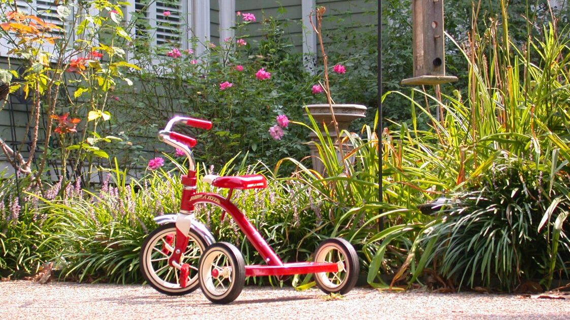 A red tricycle outside in front of a home flower bed on a sunny day