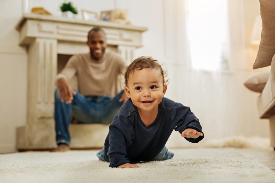 Happy father laughing and watching his cheerful young son crawling on the floor.