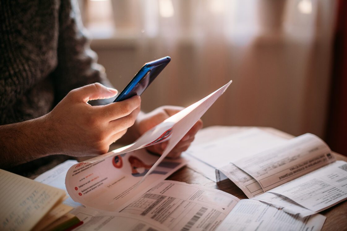 A man pays utility bills online using a QR code in an app using a smartphone in the kitchen. Close-up of hands, phone, and bills.