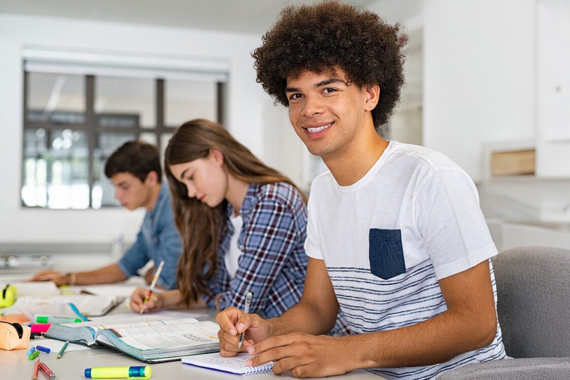 A group of teenagers sitting at a table in school which one male student looking into the camera.