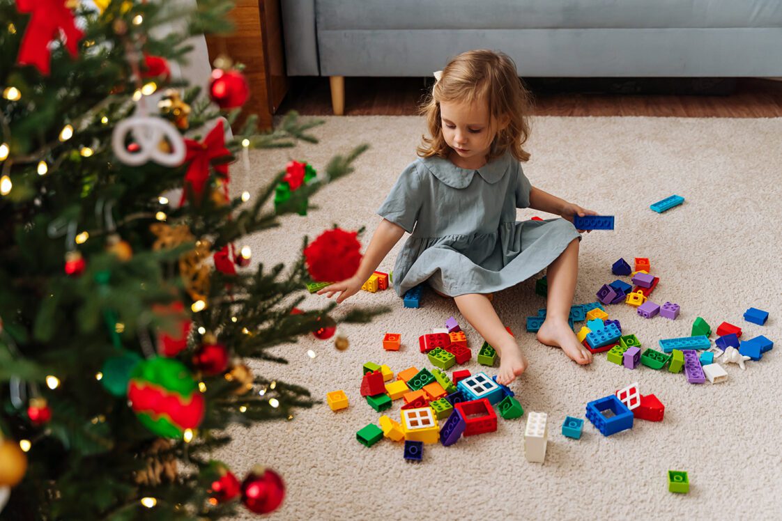 A little girl playing with colorful building blocks next to the Christmas tree.