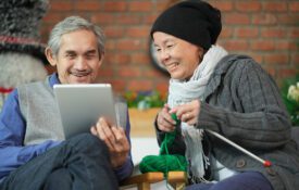 An older couple sitting on the couch during the winter. The man is holding a tablet and the woman is knitting.