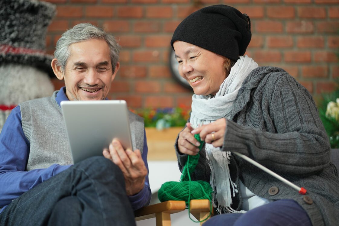An older couple sitting on the couch during the winter. The man is holding a tablet and the woman is knitting.