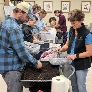 A group of Third Thursday attendees of all ages gather around a speaker to learn hands on worm composting.