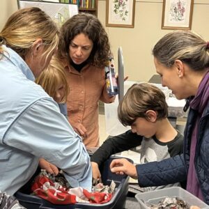 A group of Third Thursday attendees of all ages gather around a speaker to learn hands on worm composting.