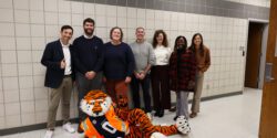 A group of Extension and Auburn University personnel with the Aubie the mascot, who is laying down on the floor with an orange construction vest and hard hat.