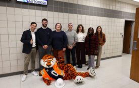 A group of Extension and Auburn University personnel with the Aubie the mascot, who is laying down on the floor with an orange construction vest and hard hat.