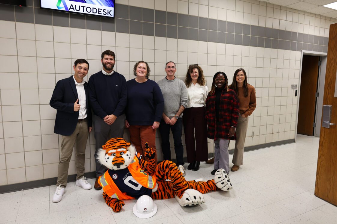 A group of Extension and Auburn University personnel with the Aubie the mascot, who is laying down on the floor with an orange construction vest and hard hat.