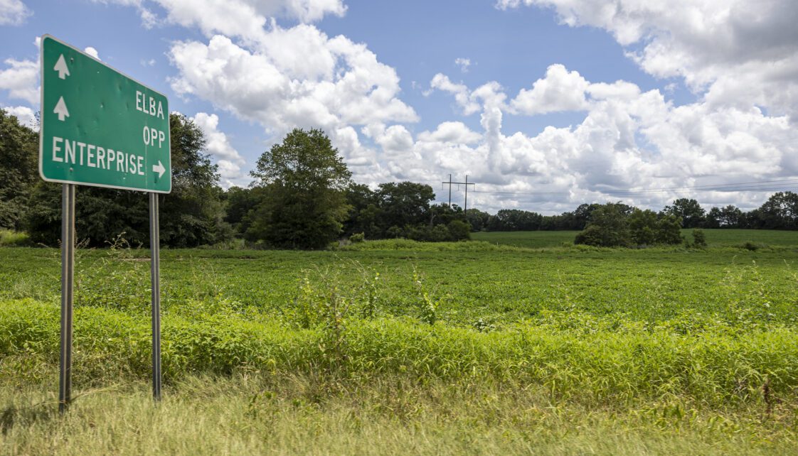 A green road sign in a rural area showing directional arrows to Elba, Opp, and Enterprise, Alabama.