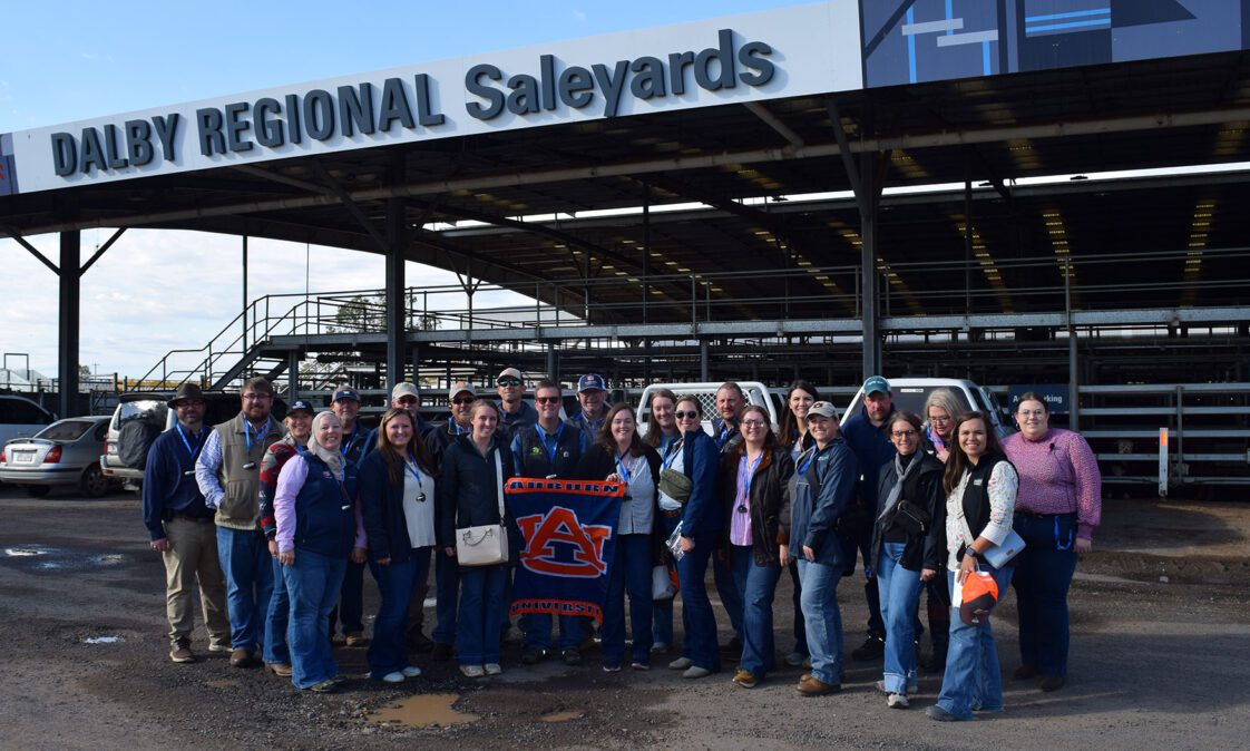 A group of approximately 30 people posing for a photo outside a saleyard facility in Australia.