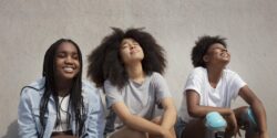 Three Black teenagers leaned up against a cement wall and looking up toward the sun with their eyes closed.