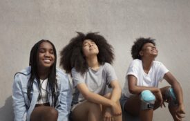 Three Black teenagers leaned up against a cement wall and looking up toward the sun with their eyes closed.