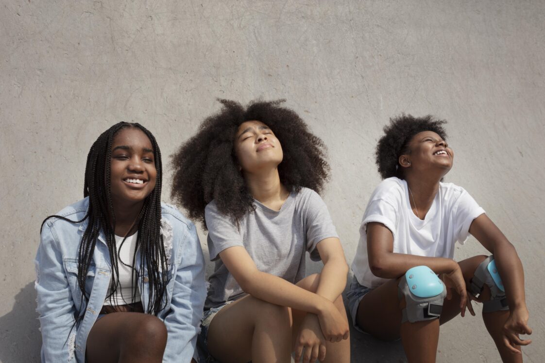Three Black teenagers leaned up against a cement wall and looking up toward the sun with their eyes closed.