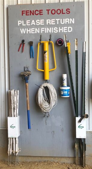 A wall with fence tools hanging on nails, including posts, a post driver, wire cutters, etc.