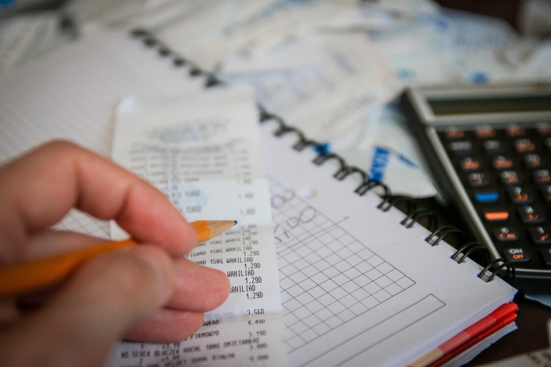 A person's hand holding a pencil over papers and a receipt while they are determining a budget with a calculator.
