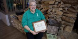Betty Gottler in a green shirt sitting on a fireplace hearth with her National 4-H Hall of Fame certificate, a photo of her parents and her parents' 4-H project books.