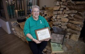 Betty Gottler in a green shirt sitting on a fireplace hearth with her National 4-H Hall of Fame certificate, a photo of her parents and her parents' 4-H project books.