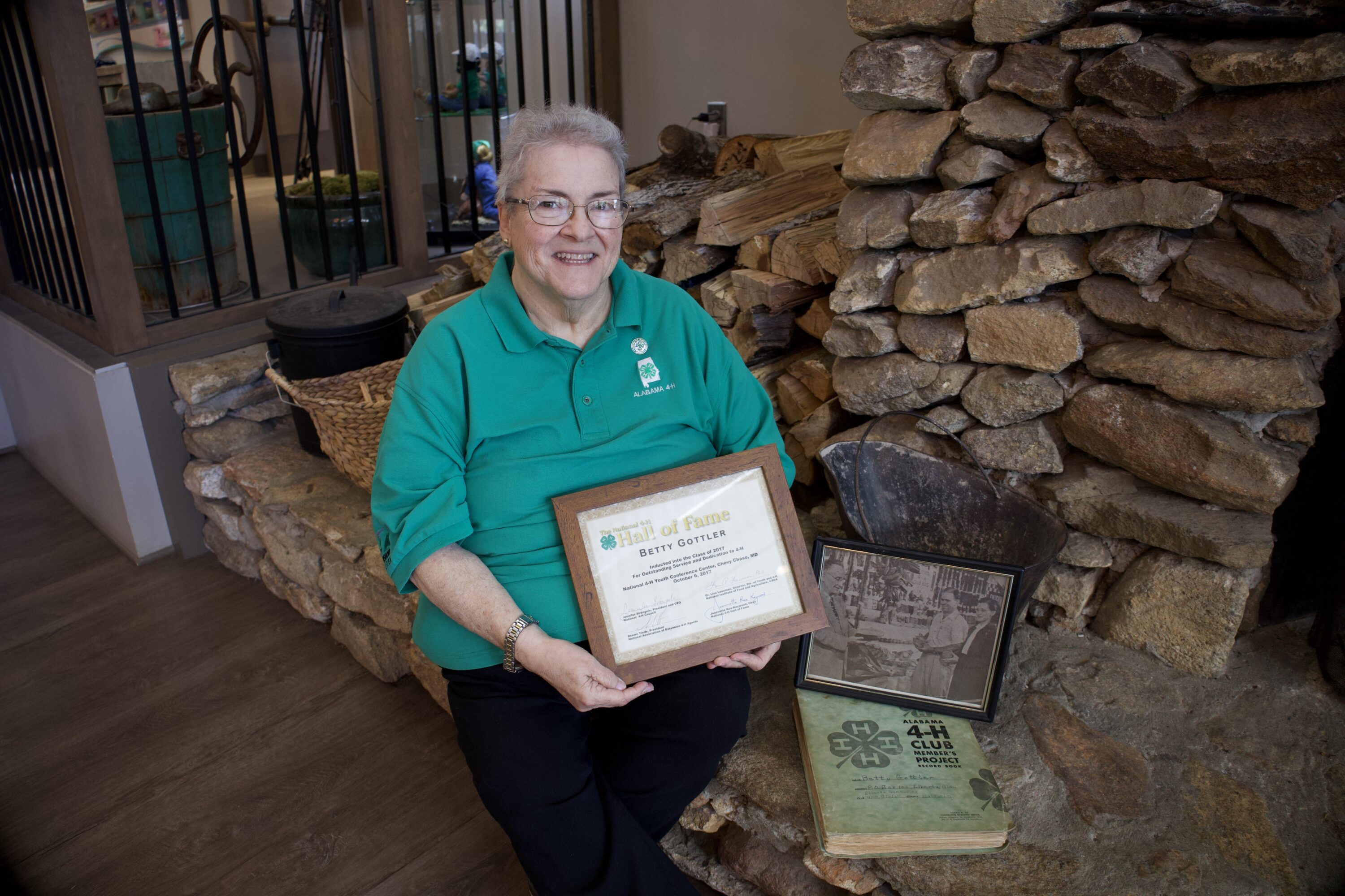 Betty Gottler in a green shirt sitting on a fireplace hearth with her National 4-H Hall of Fame certificate, a photo of her parents and her parents' 4-H project books.