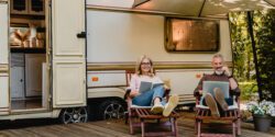 A man and woman sitting in lawn chairs under a shade umbrella in front of of a Mobile Home RV