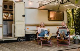 A man and woman sitting in lawn chairs under a shade umbrella in front of of a Mobile Home RV