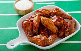 Chicken wings glazed with honey in a whit platter on a table with a tablecloth that looks like a football field. A cup of sauce in background.