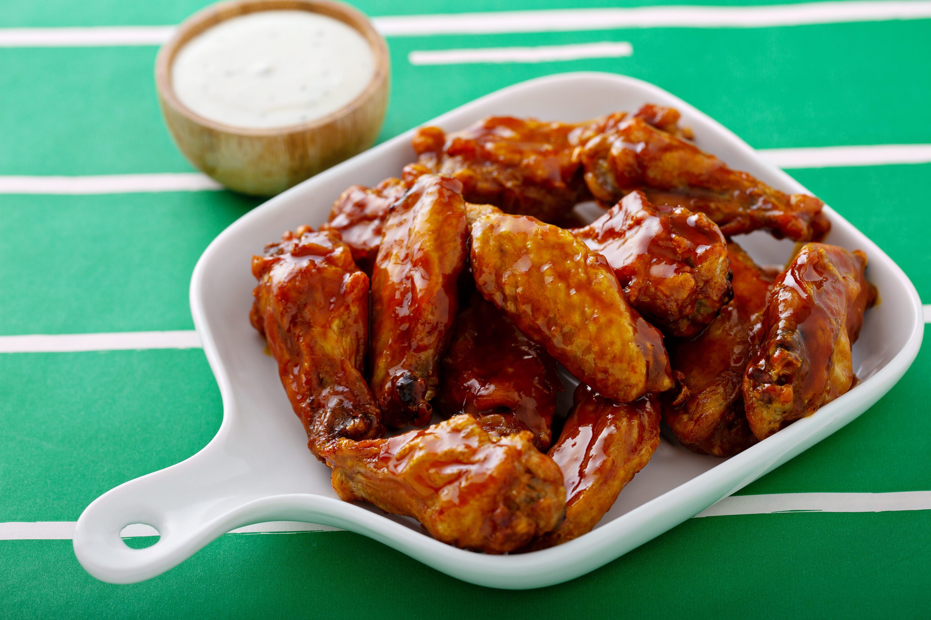 Chicken wings glazed with honey in a whit platter on a table with a tablecloth that looks like a football field. A cup of sauce in background.