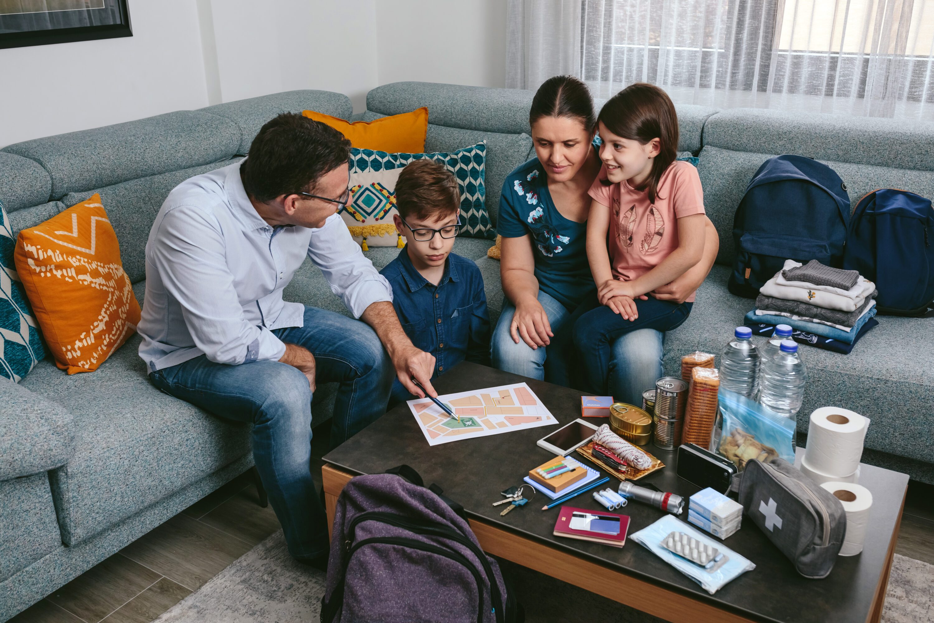 A family sitting on a couch around a coffee table that is stocked with emergency supplies. They are reviewing an emerency plan.