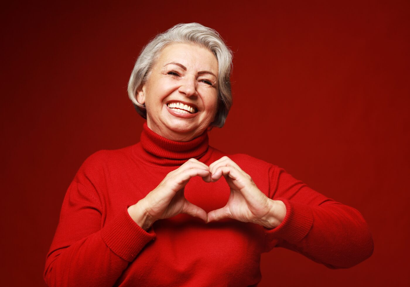 Older gray-haired woman wearing red sweater, smiling and making a heart symbol with her hands. For American Heart Month.