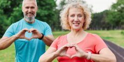 Smiling, active, mature, middle-aged couple posing together. Both are making heart symbols with their hands. There is a paved walking path behind them. For American Heart Month.