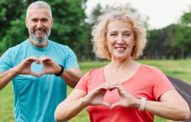 Smiling, active, mature, middle-aged couple posing together. Both are making heart symbols with their hands. There is a paved walking path behind them. For American Heart Month.