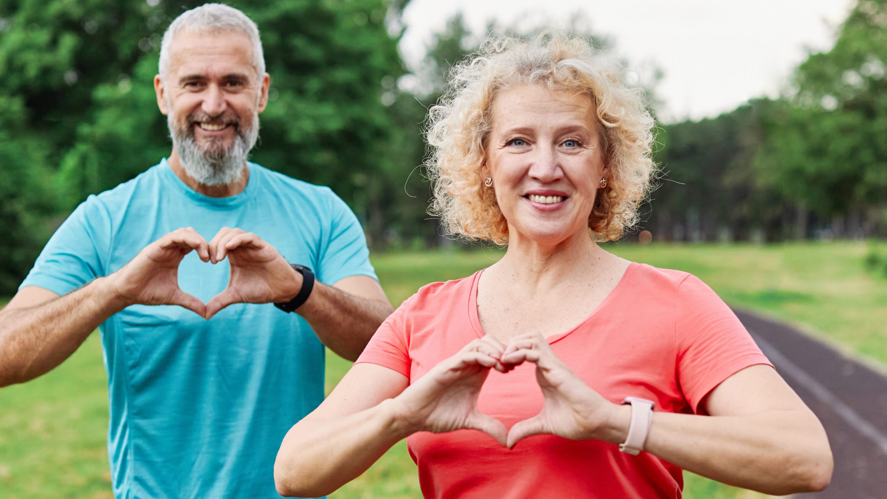 Smiling, active, mature, middle-aged couple posing together. Both are making heart symbols with their hands. There is a paved walking path behind them. For American Heart Month.