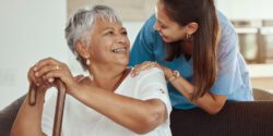 Happy, relax and senior woman with caregiver smile while sitting on a living room sofa in a nursing home. Support, help and professional nurse or healthcare worker helping elderly lady or patient.